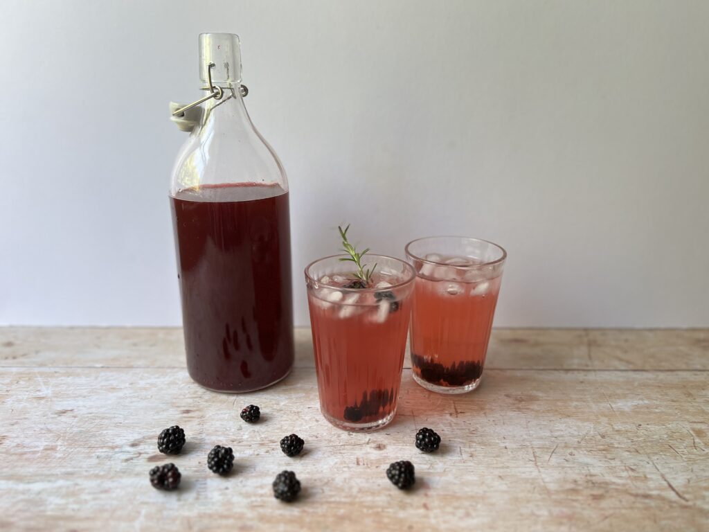 bottle of home made bramble shrub with two glasses of shrub drink